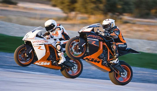 Deux motos de sport KTM en pleine course sur un circuit, dont l'une effectue un wheelie, illustrant la vitesse, la maîtrise et l'action propre aux sports mécaniques de compétition.