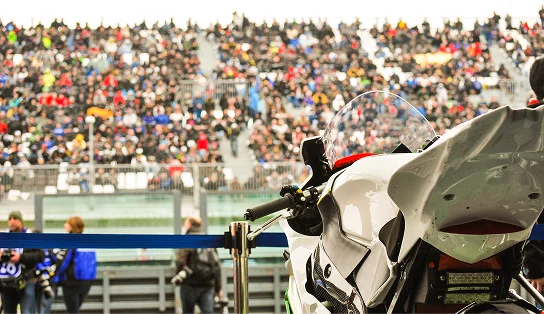 Une moto de sport garée près de la voie des stands, avec une foule nombreuse dans les tribunes du stade, illustrant l'ambiance d'une course de moto en direct.