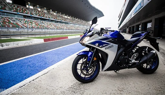 Une moto de sport Yamaha bleue garée dans la voie des stands d'un circuit automobile, avec les tribunes et la piste en arrière-plan, illustrant l'univers du sport automobile professionnel.